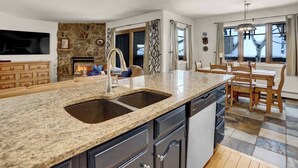 Eine Küche mit Granit Arbeitsplatten, einem Geschirrspüler, einer Spüle, einer Wand mit Steinverkleidung, einem Kamin und einem Essbereich mit Holztisch und -stühlen.