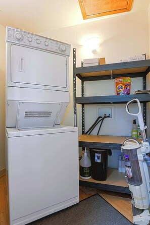 A compact laundry area with a stacked washer and dryer, a shelf with cleaning supplies, and a small table with a lamp. - Werner Meadows Penthouse (Steamboat Springs)