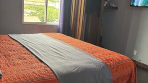 Ein Schlafzimmer mit einem Bett, einem Fenster mit Vorhängen, einem Fernseher und einem an der Wand befestigten Regal.