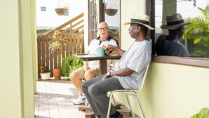 Deux hommes sont assis à une table sur une terrasse en bois, entourée de plantes en pot et d’un panier suspendu.