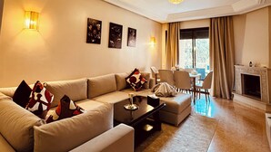 A spacious living room with a large beige sofa, a coffee table, and a fireplace. There are wall-mounted lights and a chandelier. - Marrakech Oasis 2-bedroom apartment with direct garden and pool access with AC (Marrakech)