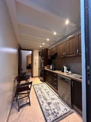 A compact kitchen with dark wooden cabinets, a built-in oven, a microwave, and a gas stove. There is a small dining area with a table and chairs. - Marrakech Oasis 2-bedroom apartment with direct garden and pool access with AC (Marrakech)