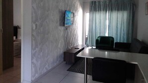 A modern living room with a white marble wall, a flat-screen TV mounted above a dark wooden coffee table, a green armchair, and a large window with light blue curtains. - Wonderful apartment in peaceful Midrand perfect for your stay (Midrand)