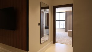 A hallway with a flat-screen TV mounted on the wall, a large mirror, and a door leading to a room with a carpeted floor and a window with curtains.