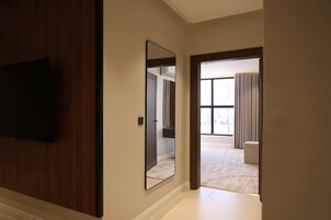Un pasillo con una televisión de pantalla plana montada en la pared, un espejo grande y una puerta que da a una habitación con piso alfombrado y una ventana con cortinas.