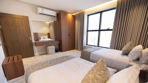 A hotel room with two beds, a wooden wardrobe, a bathroom with a mirror, and a large window with curtains. - Royal Edition Hotel Apartments (Buraydah)