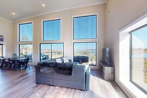 A spacious living area with large windows, a grey sofa, a dining table with chairs, and a flat-screen TV. - Lakefront Oasis w/ Water Toys! Breckenridge Home (Breckenridge)