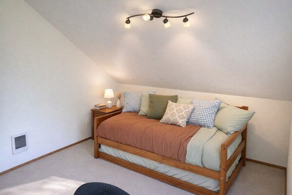 A bedroom with a sloped ceiling, a wooden bed, a bedside table with a lamp, and a wall-mounted light fixture. - Restorative Island Retreat - Beach Access and Private Half-Acre (Coupeville)