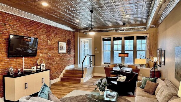 Una sala de estar con pared de ladrillo, piso de madera, un sofá, una mesa de centro, un televisor y una escalera que lleva a otra habitación.