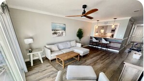 A modern living room with a sofa, ottoman, and coffee table, a dining area with chairs, and a kitchen in the background. - 🌴 Waterfront-Beach-Swimming-Fishing-Dock-Double sized private Balcony (Mary Esther)