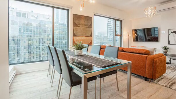 A modern living room with a glass dining table, orange sofa, and large windows offering city views. - Free Indoor Parking Old Montreal 4 (Montréal)