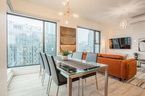 A modern living room with a glass dining table, orange sofa, and large windows offering city views. - Free Indoor Parking in Old Montreal 4 (Montréal)