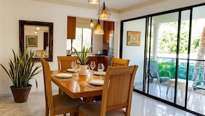 A dining area with a wooden table, chairs, and hanging pendant lights. There is a potted plant on the floor and a large window with a view of the outdoors. - Stylish 2BR in Sosua with Pool, Bbq, Fast Internet & Back up Generator (Sosúa)