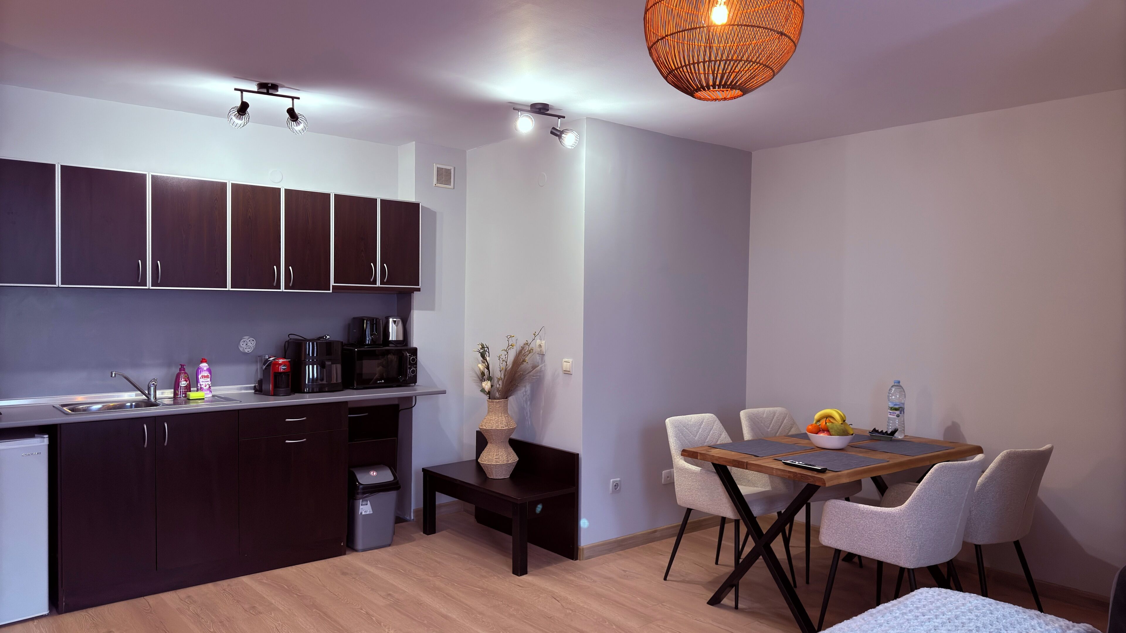 A modern kitchen with dark cabinets, a dining area with a wooden table and white chairs, and a hanging light fixture.
