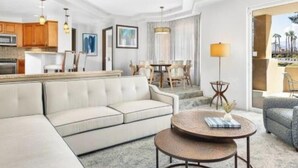 A modern hotel room with a living area featuring a sofa, a round coffee table, and a dining table with chairs. There is a kitchen area with wooden cabinets and a microwave. - JW Marriott's Desert Springs Villas I - Access to Marriott Spa, Sleeps 6-8 (Palm Desert)