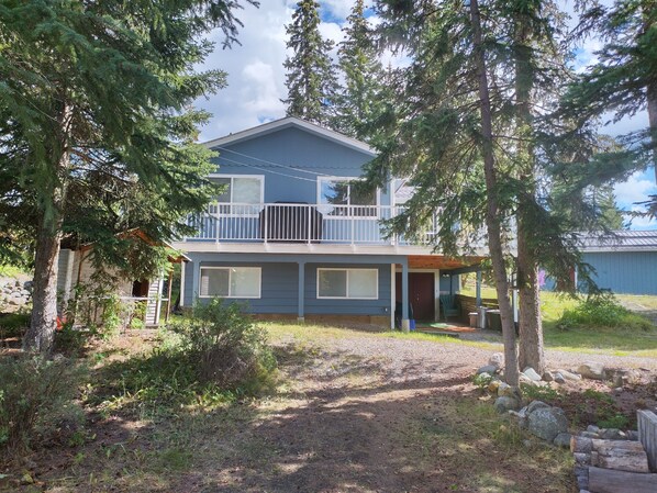 A two-story house with a balcony, surrounded by trees and a gravel driveway.