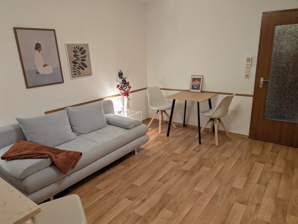 A modern living room with a sofa, a dining table with chairs, a vase with flowers, and framed artwork on the wall.