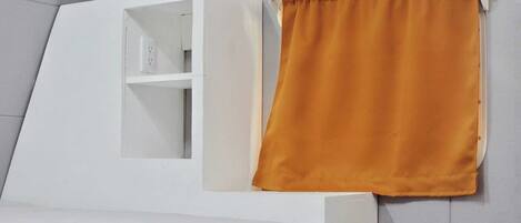Una cama blanca con cabecera, una mesita de noche blanca y una cortina color mostaza.