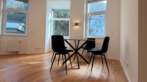 Une pièce spacieuse dotée d’un plancher en bois, d’un luminaire rond au plafond et d’une table entourée de quatre chaises. Trois fenêtres laissent entrer la lumière naturelle et offrent une vue sur l’extérieur.