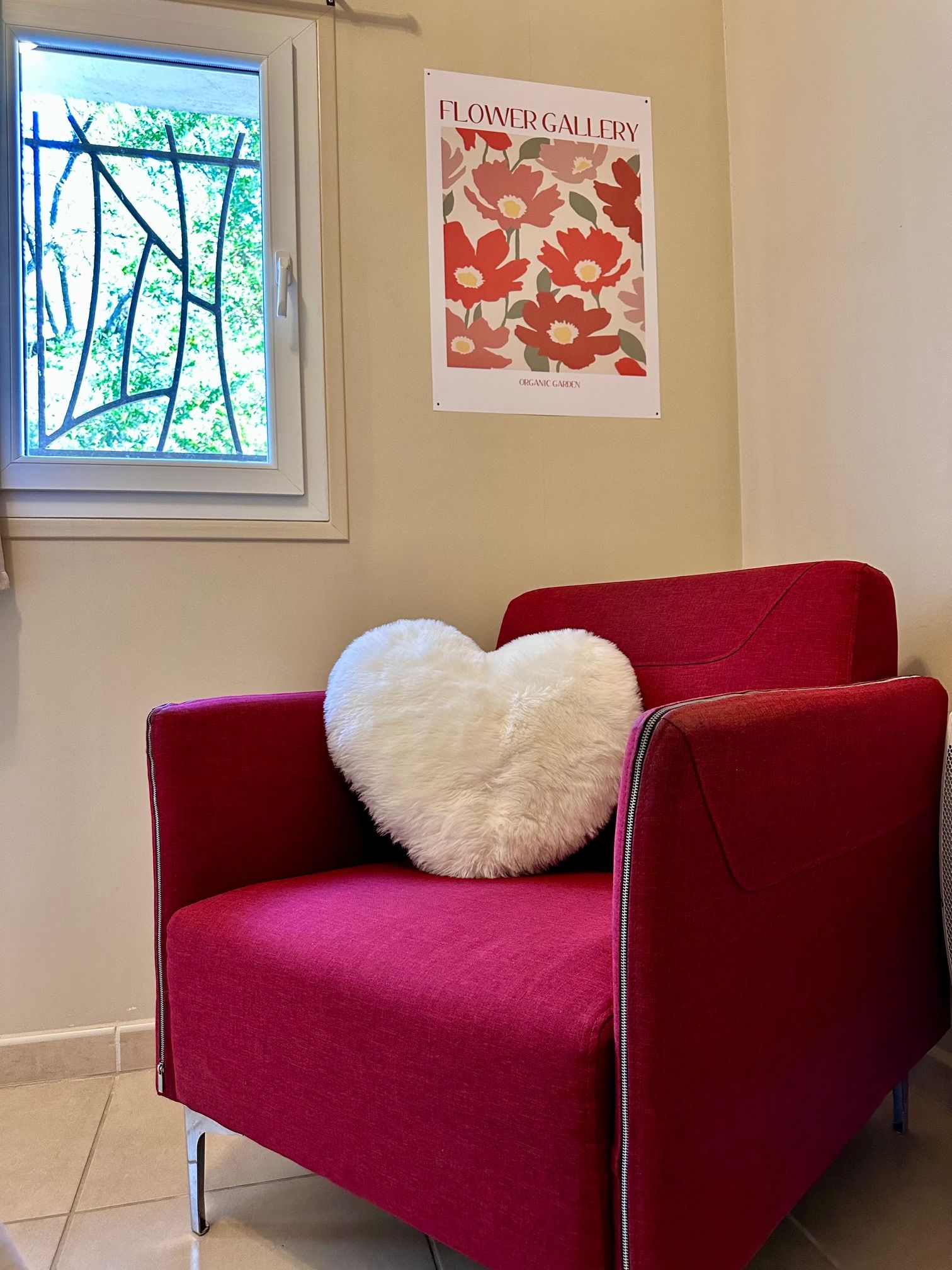A red chair with a white heart-shaped pillow, a window with a view of greenery, and a poster on the wall titled "Flower Gallery".