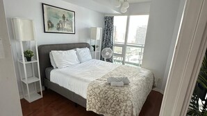 Ein Schlafzimmer mit einem Bett, einer weißen Regaleinrichtung, einem Gemälde und einem Fenster mit Blick auf Gebäude.