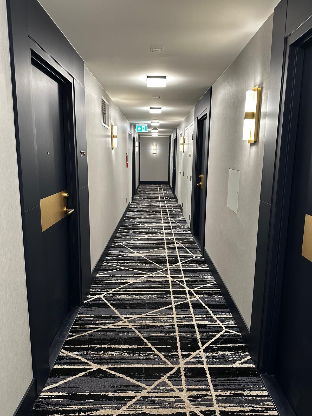 A hotel corridor with doors on both sides, a patterned carpet, and wall-mounted light fixtures.