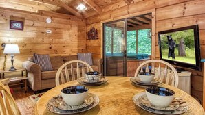 Interior acogedor de una cabaña con paredes de madera, una mesa de comedor dispuesta con fuentes, un sofá y un televisor mostrando un oso.