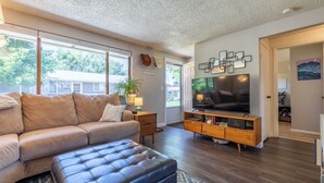 A modern living room with a leather sofa, a tufted ottoman, a wooden TV stand, and a large flat-screen TV. - 8th St Retreat: Loveland Comfort (Loveland)