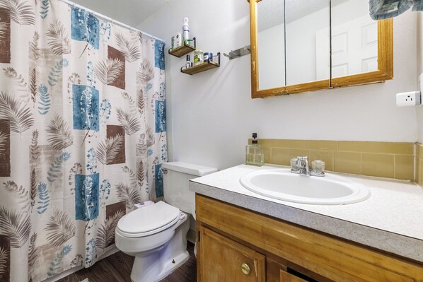 A bathroom with a toilet, a sink, a mirror, and a shower curtain with a floral pattern. - 8th St Retreat: Loveland Comfort (Loveland)
