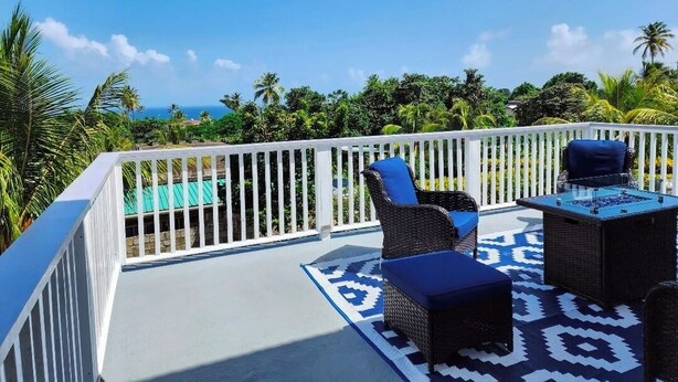 A balcony with wicker furniture and a patterned rug, overlooking a pool and lush greenery. - Entire rental unit in Calibishie, Dominica (Calibishie)