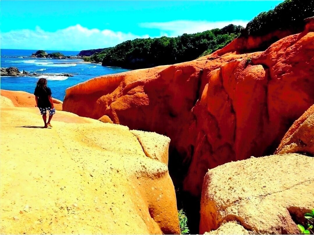 Eine Person steht an einem sandigen Strand mit roten Felsformationen und dem Meer im Hintergrund.