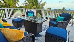 A covered outdoor seating area with wicker furniture and cushions, a glass-top table, and a view of the ocean and palm trees. - Entire rental unit in Calibishie, Dominica (Calibishie)