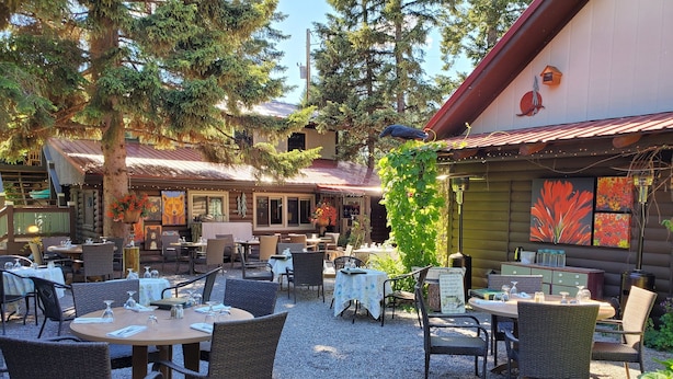 An outdoor dining area with tables and chairs, surrounded by trees and a building with a red roof. - Laughing Horse Lodge (Bigfork)
