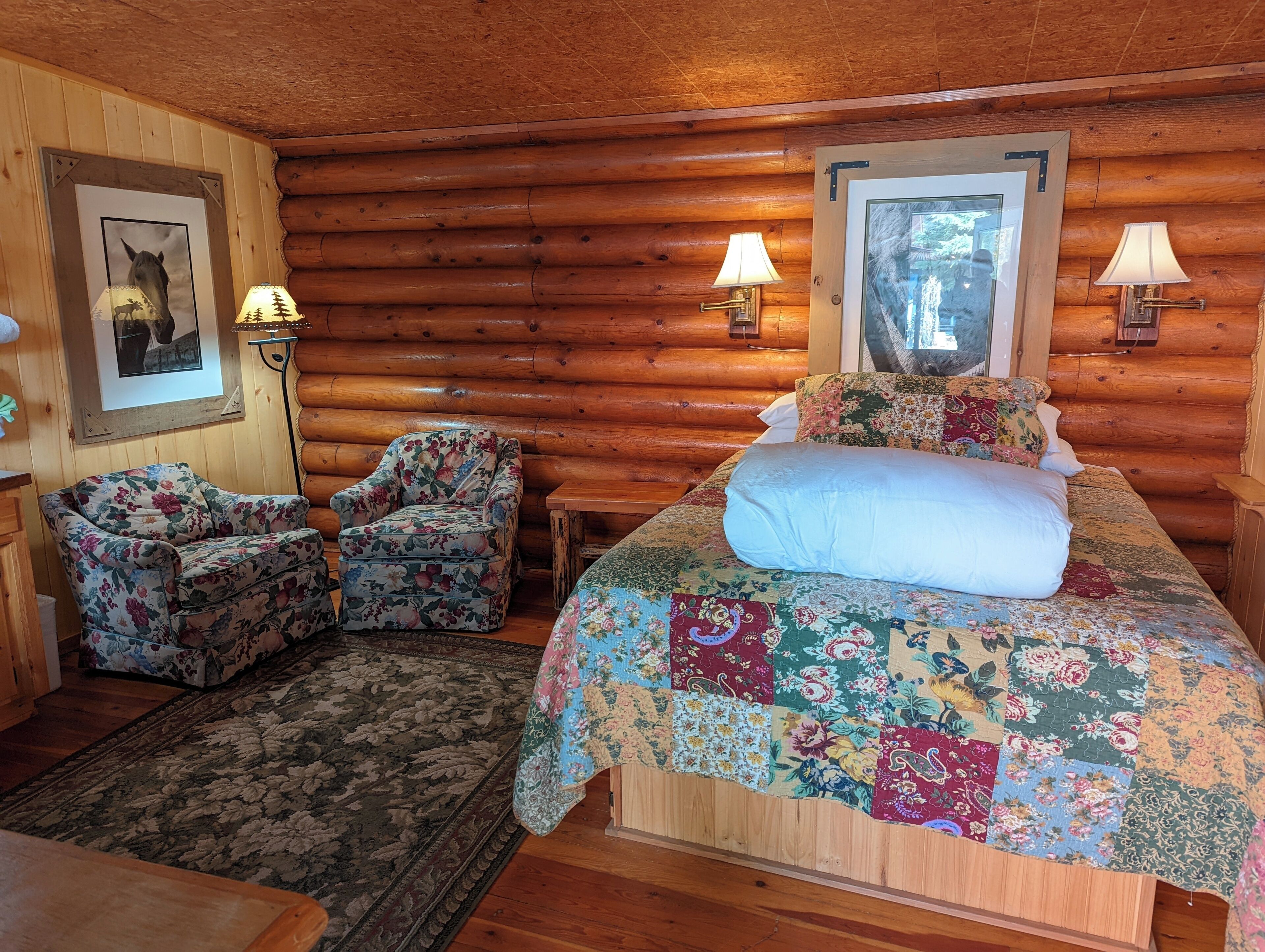 A log cabin room with a bed, two chairs, a rug, and two wall-mounted lamps.