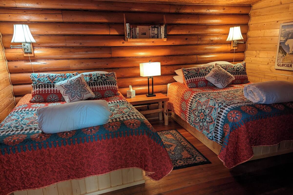Acogedora habitación tipo cabaña de madera con dos camas, cada una con ropa de cama y almohadas estampadas, un pequeño mesita de noche y lámparas de pared.