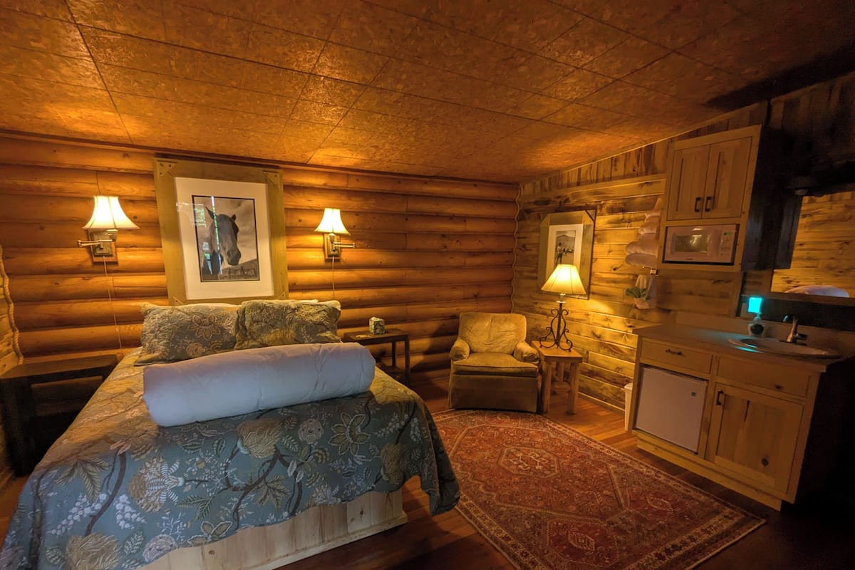 Un dormitorio tipo cabaña de troncos con cama, dos sillones, una mesita, una lámpara, un cuadro en la pared y una pequeña cocina.