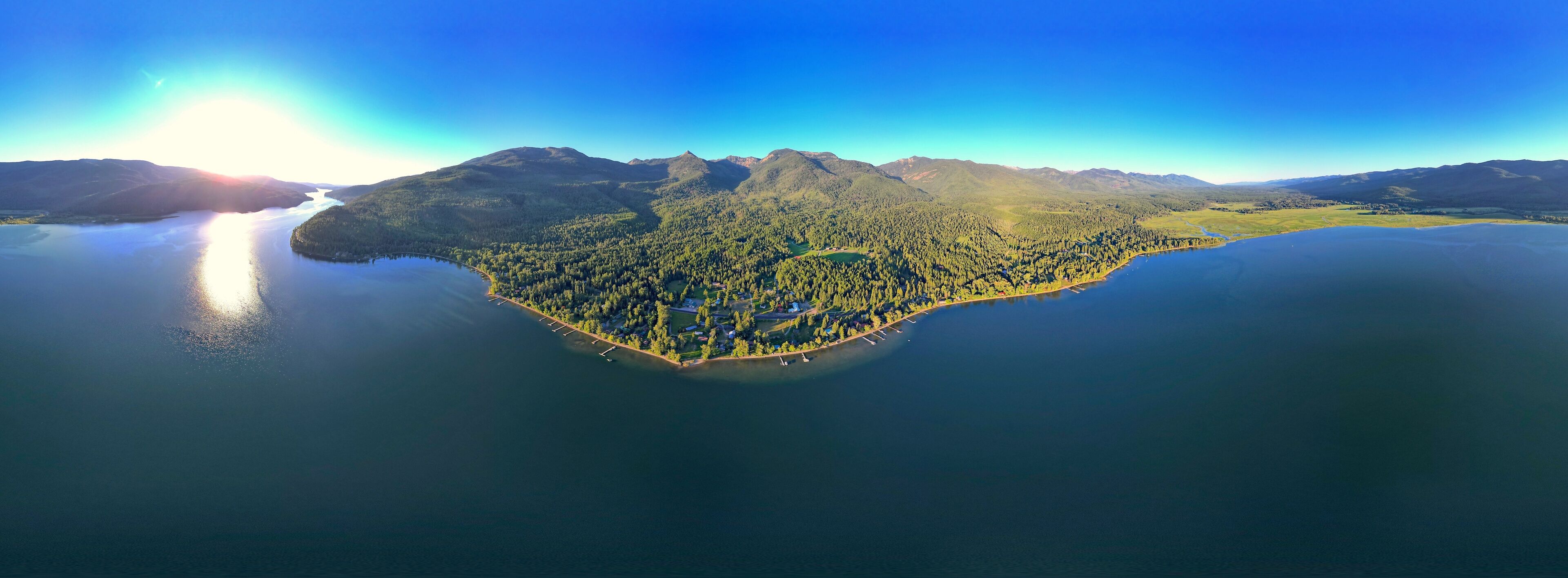 An aerial view of a lakeside island with dense forestation, a marina, and mountainous terrain in the background.