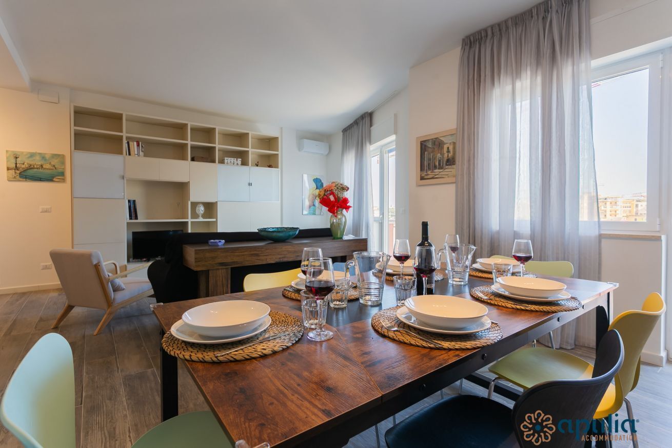 Un coin repas avec une table en bois dressée pour quatre personnes, entourée de chaises. On aperçoit, au fond, un espace cuisine avec un comptoir et un évier.