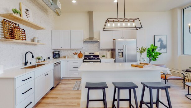 Une cuisine moderne avec des meubles blancs, un îlot central doté de tabourets, un réfrigérateur en acier inoxydable et un luminaire suspendu.