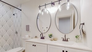 Une salle de bain avec un lavabo blanc, deux miroirs, des toilettes et une douche dont le carrelage présente un motif géométrique.