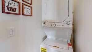 Une machine à laver blanche, dotée de boutons de commande et d’une étiquette. Des panneaux encadrés, avec des mots liés au lavage du linge, sont accrochés au mur.