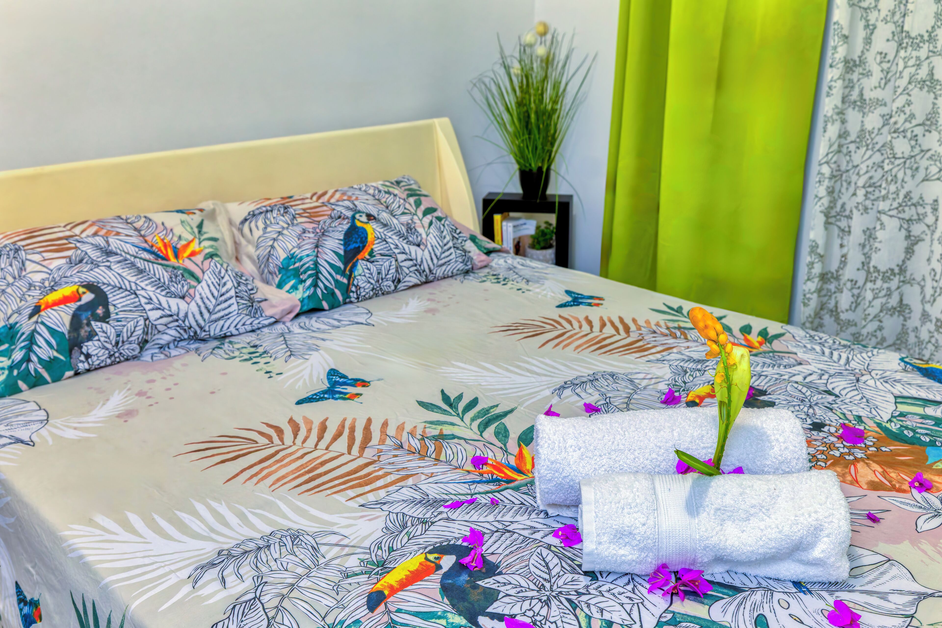 Un lit recouvert d’une couvre-lit à motifs floraux et d’oiseaux, une petite plante posée sur une table de chevet et un rideau vert.