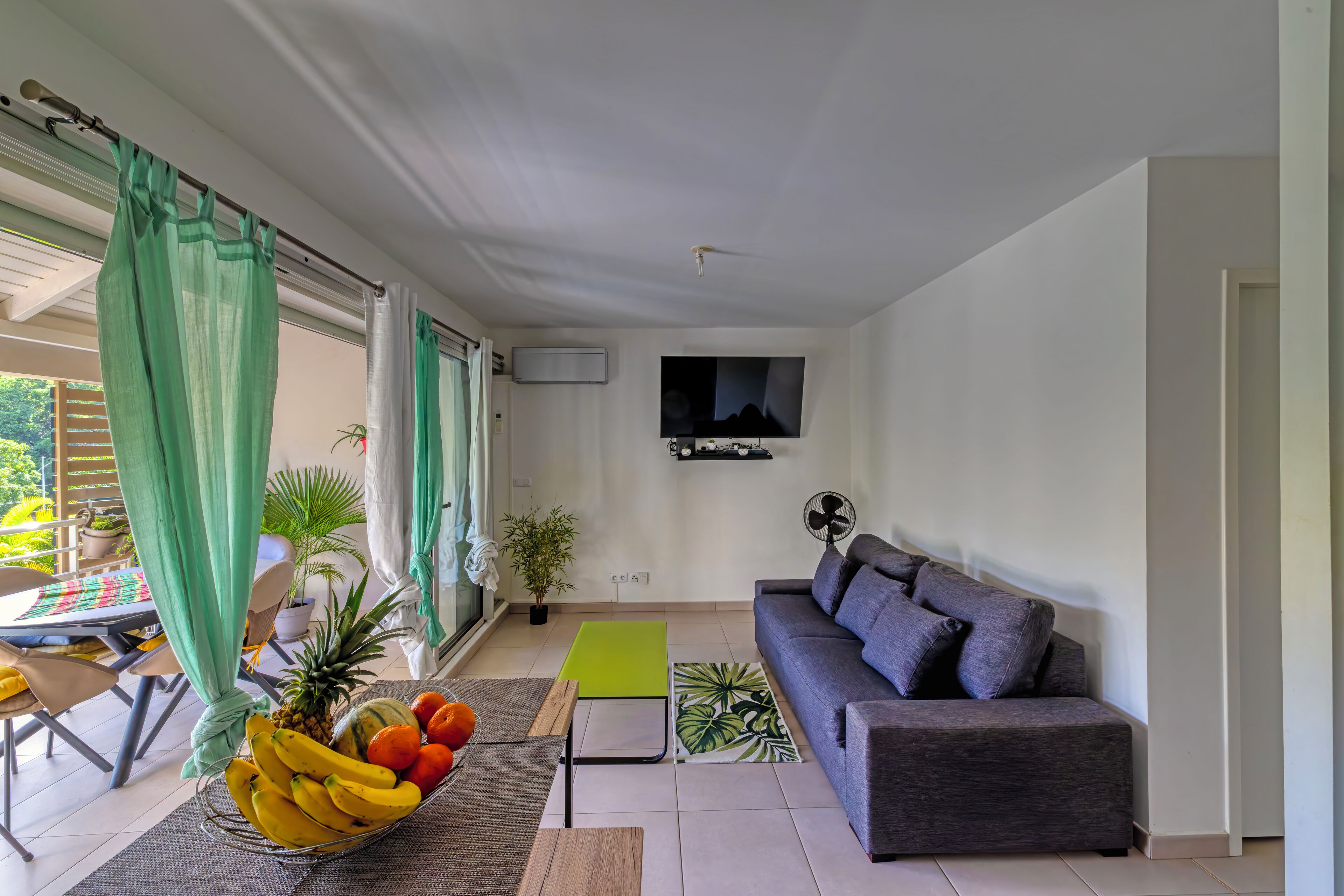 Un salon moderne avec un canapé gris, un téléviseur à écran plat fixé au mur, un coin repas avec une table et des chaises, et un panier de fruits posé sur le sol.