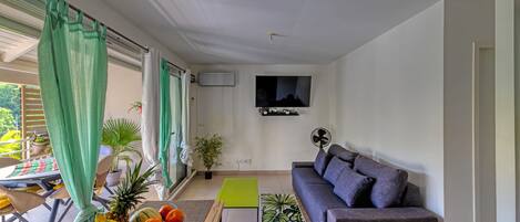 Un salon moderne avec un canapé gris, un téléviseur à écran plat fixé au mur, un coin repas avec une table et des chaises, et un panier de fruits posé sur le sol.