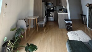 Eine moderne Küche mit Edelstahlgeräten, ein Essbereich mit einem kleinen runden Tisch und Stühlen sowie ein Wohnbereich mit einer Couch und einer großen Pflanze.