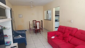 A living room with a red sofa, a blue sofa, a dining table with chairs, and a kitchen area in the background. - Cozy and very well-located apartment in a condominium. (Praia Grande)