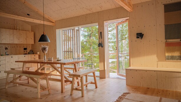 Un intérieur en bois avec une table à manger et des bancs, un luminaire suspendu et une vue à travers une porte vitrée.
