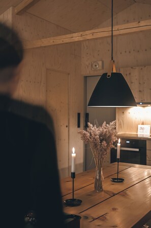 Une table en bois ornée de bougies et d’un vase de fleurs séchées, une personne au premier plan et une lampe suspendue.