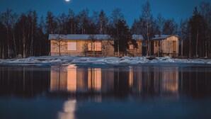 Une cabane dotée d’une grande fenêtre et d’une plus petite, entourée d’un paysage enneigé et d’arbres, avec un lac paisible qui reflète la scène.