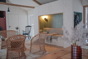 A cozy room with a wooden floor, a sofa with cushions, wicker chairs, and a table with a vase of dried flowers.